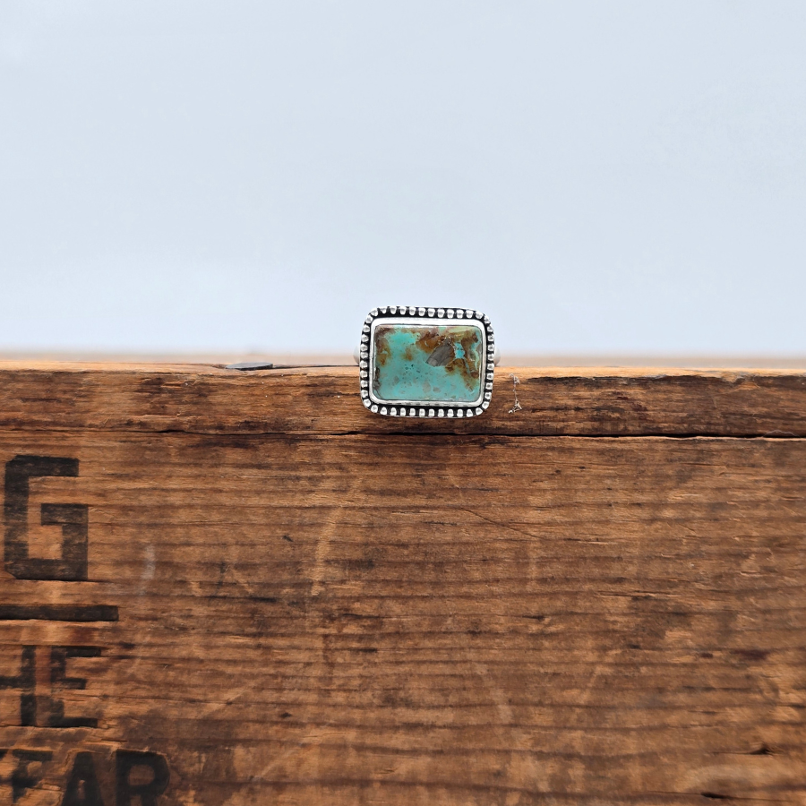 A silver-toned ring with a rectangular turquoise stone, displayed on a rustic wooden surface against a pale blue sky background.